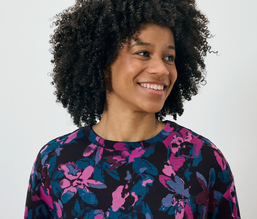 Porträt einer lächelnden Frau mit Afro, die ein schwarzes Oberteil mit rosa-blauem Blumenmuster trägt.