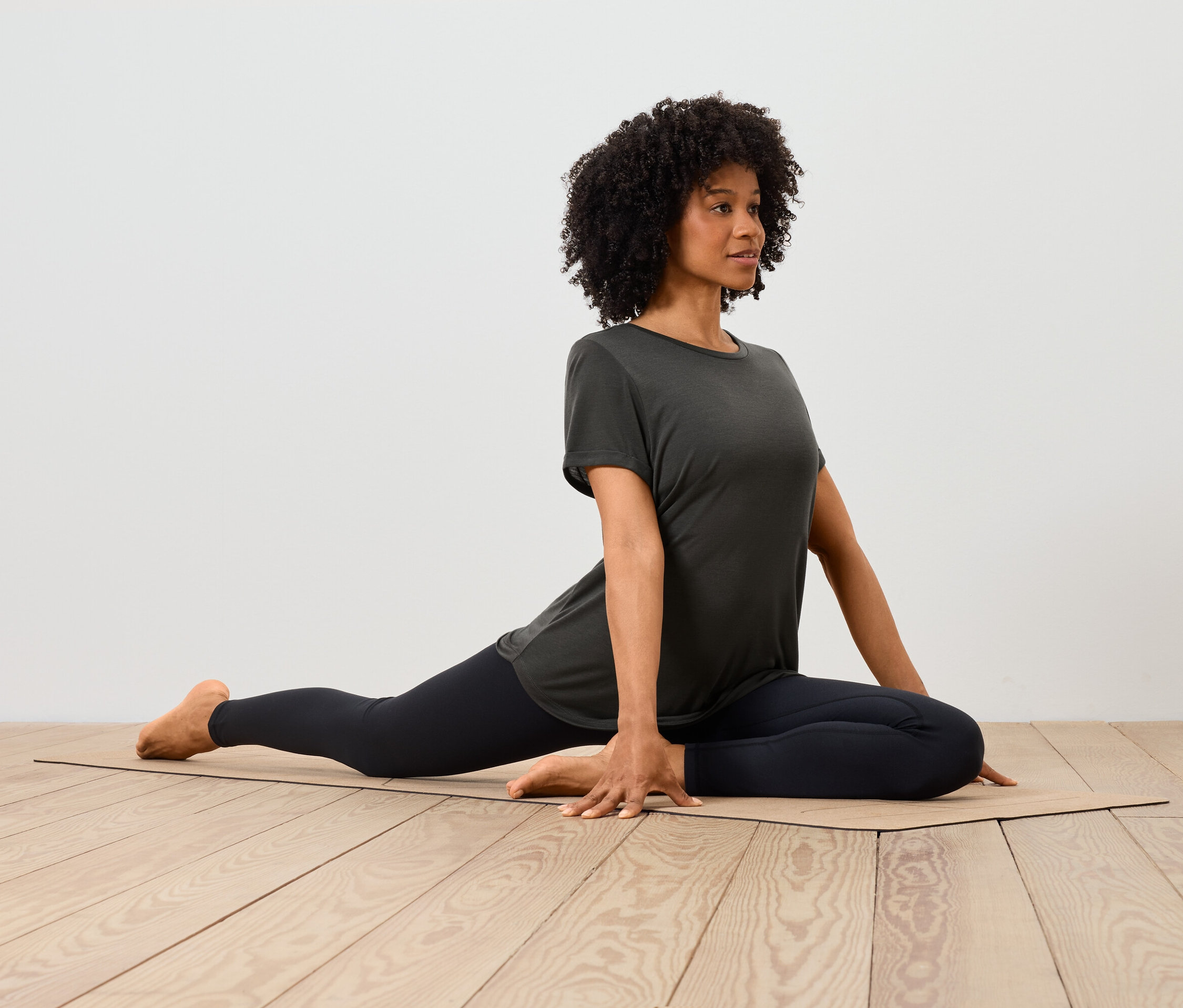 Eine afroamerikanische Frau in T-Shirt und Leggings dehnt sich auf einer Matte auf einem Holzboden.