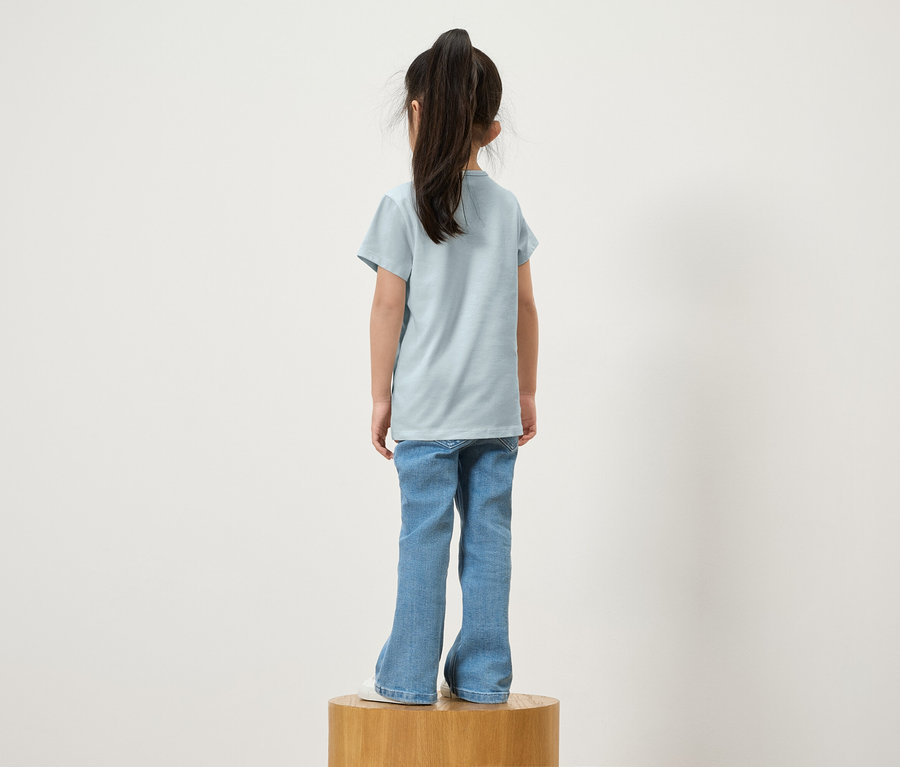 Ein Mädchen mit Pferdeschwanz steht von hinten auf einem Holzsockel, trägt ein blaues T-Shirt und Jeans.