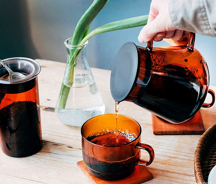 Eine Hand gießt Tee aus einem bernsteinfarbenen HARIO »Colors« Tee- und Kaffee-Set in eine Tasse.