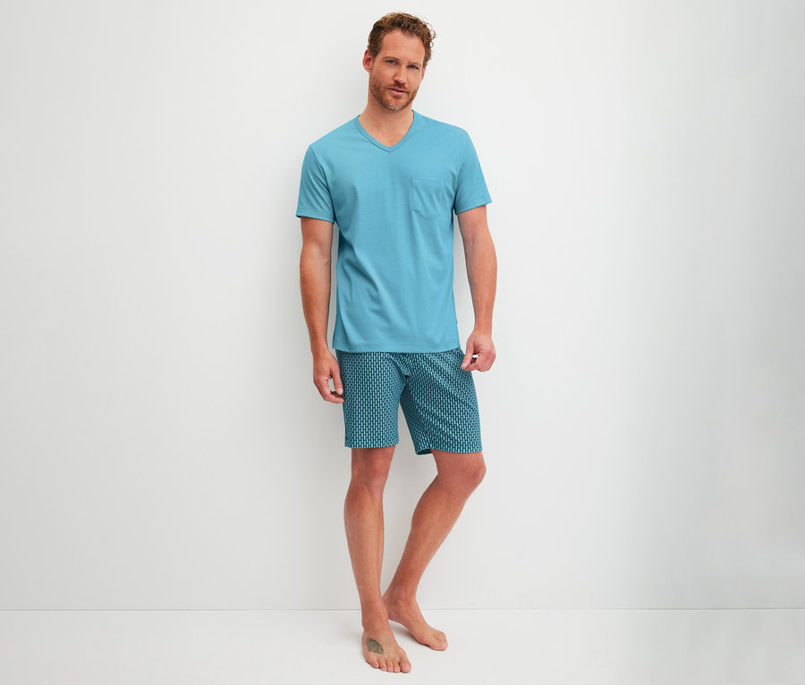 Mann steht barfuß in hellblauem T-Shirt und gemusterten Shorts vor einer weißen Wand.