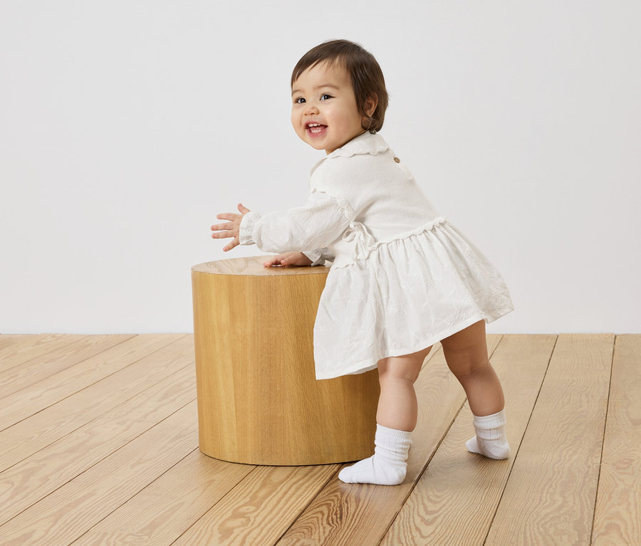 Ein lächelndes Baby in einem weißen Kleid steht neben einem kleinen Holzhocker auf einem Holzboden.