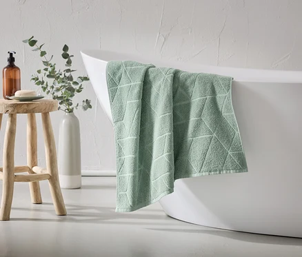 Ein mintgrünes Handtuch mit geometrischem Muster hängt über dem Rand einer weißen Badewanne. Ein Holzhocker mit Seife und Spender steht daneben.