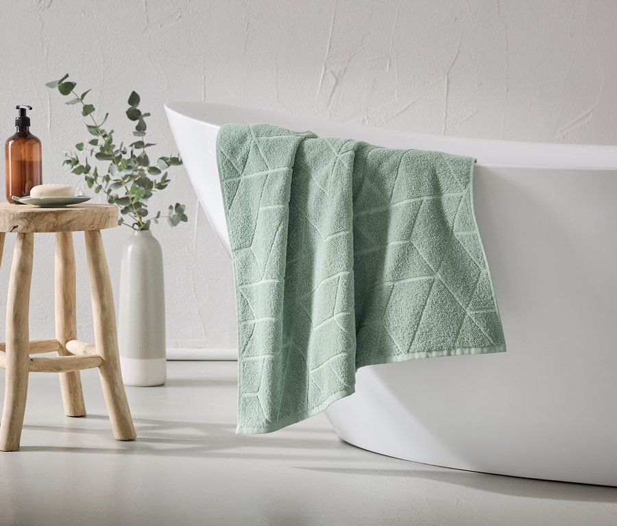 Ein mintgrünes Handtuch mit geometrischem Muster hängt über dem Rand einer weißen Badewanne. Ein Holzhocker mit Seife und Spender steht daneben.