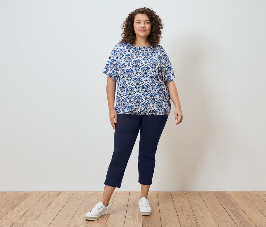 Eine Frau mit lockigem Haar steht vor einer Wand und trägt eine blau gemusterte Bluse, marineblaue Hosen und weiße Turnschuhe.