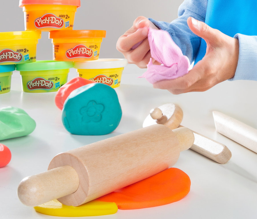 Eine Person formt rosa Play-Doh in ihren Händen, umgeben von Play-Doh-Behältern, einem Nudelholz und bunten Play-Doh-Formen.