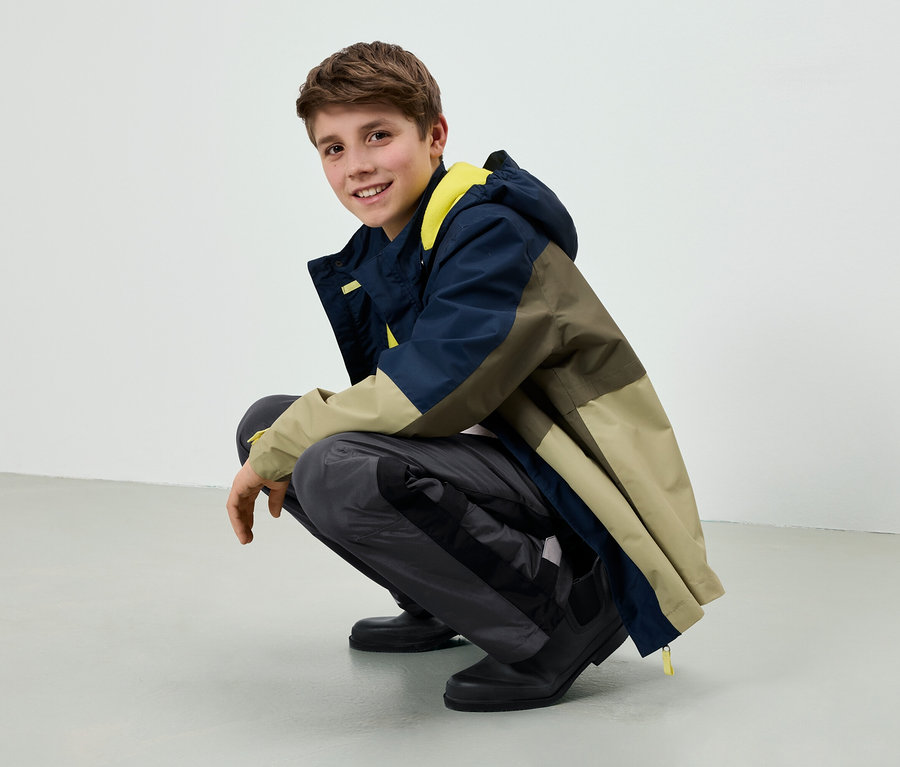 Ein Teenager hockt, lächelt und trägt eine farblich abgesetzte Jacke in Beige, Oliv und Marineblau mit gelbem Futter, graue Hose und schwarze Schuhe.