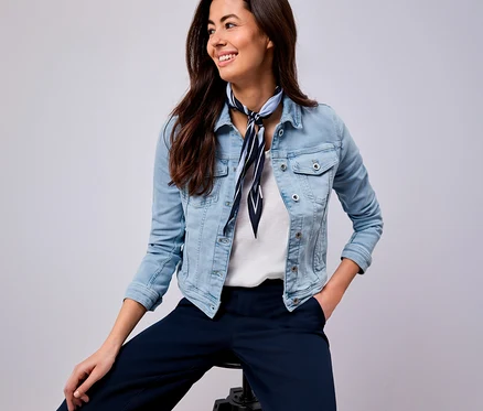 Lächelnde Frau mit blau-weiß gestreiftem Tuch, Jeansjacke und blauer Hose sitzt auf einem Hocker.