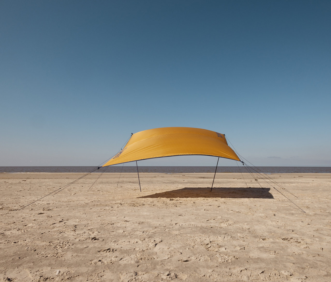 Ein gelbes Tambu »CHADA« Tarp/Sonnensegel steht am Strand.