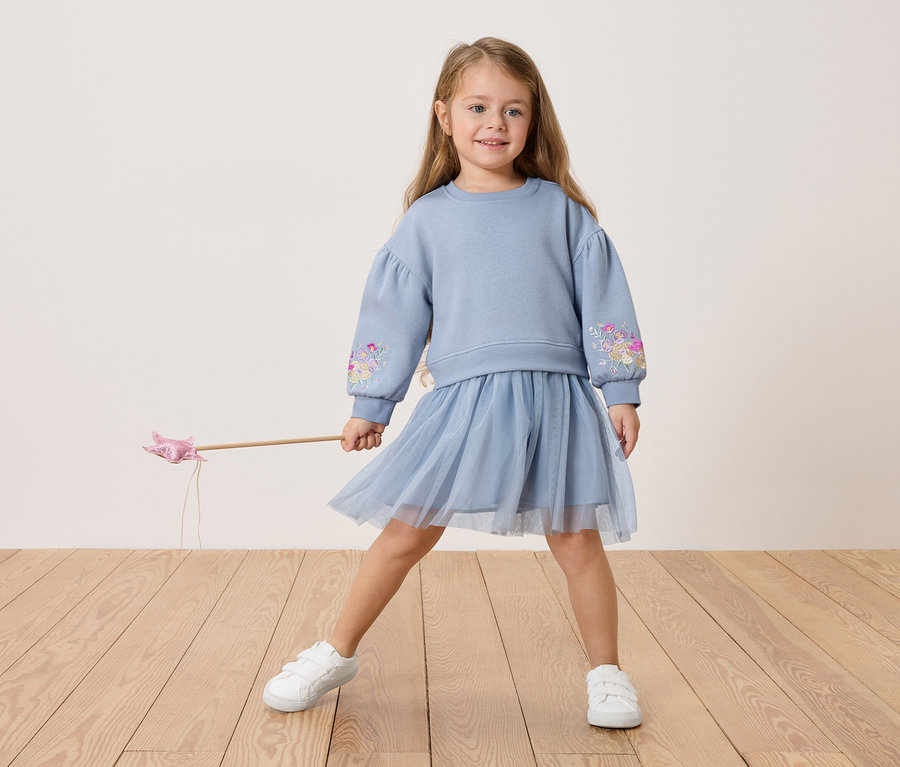 Mädchen mit blauem Pullover mit Blumenstickerei und Tüllrock, hält einen Zauberstab.