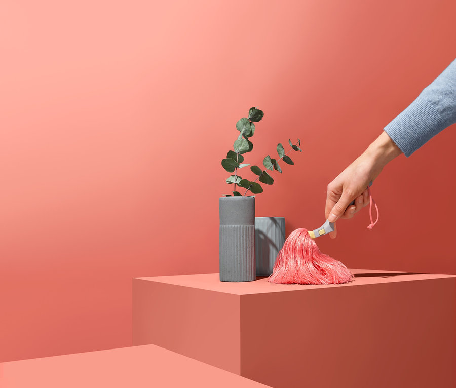 Hand hält Staubwedel mit rosa Quaste neben zwei grauen Vasen mit Pflanzen auf korallenrotem Hintergrund.