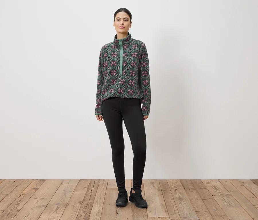Eine Frau steht vor einer Wand und trägt ein grün-pink gemustertes Fleeceoberteil, Leggings und Turnschuhe.