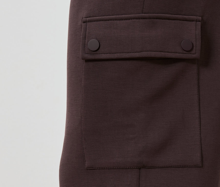 Detailansicht einer kastanienbraunen Hose mit aufgesetzter Tasche, die mit zwei runden Knöpfen geschlossen wird.