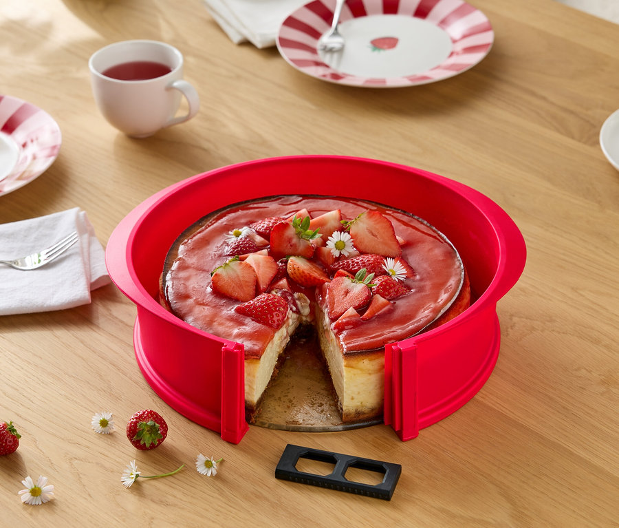 Erdbeer-Käsekuchen mit rotem Tortenring, angeschnitten, auf Holztisch mit Gänseblümchen, Gedeck und Getränken.