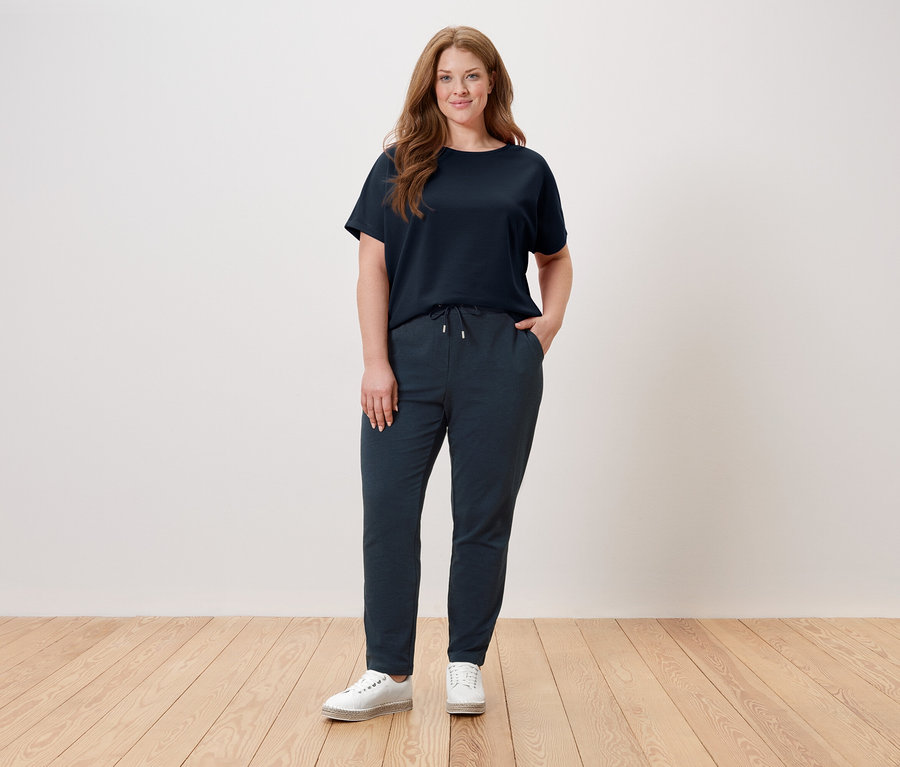 Eine Frau steht vor einer Wand und trägt ein dunkelblaues T-Shirt, eine dunkelblaue Hose und weiße Schuhe.