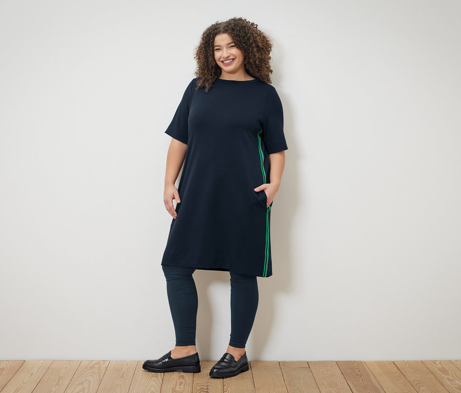 Frau mit lockigem Haar trägt ein marineblaues Kleid mit grünen Streifen, marineblaue Leggings und schwarze Slipper.