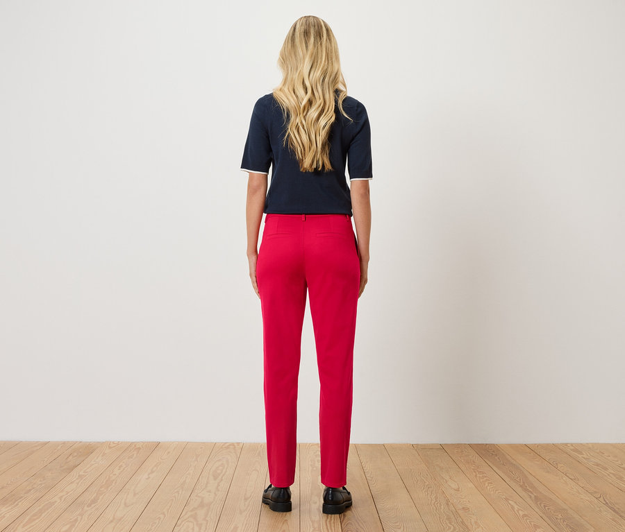 Eine Frau mit blonden, gewellten Haaren steht mit dem Rücken zur Kamera, trägt ein marineblaues Oberteil und eine rote Hose.