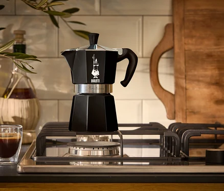 Ein schwarzer Espressokocher steht auf einem Gasherd neben einem Glas Espresso.