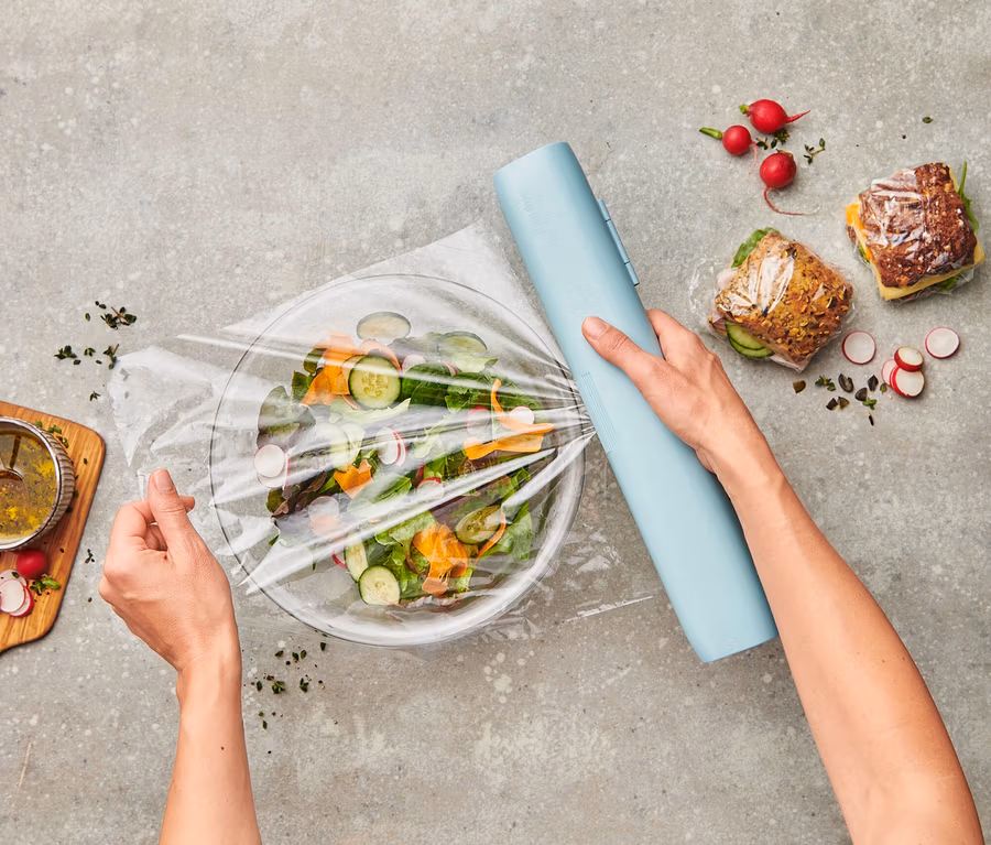 Hände verschließen Salatschüssel mit Frischhaltefolie, die von einem hellblauen Spender abgerollt wird. Daneben liegen belegte Brote und Salatdressing.