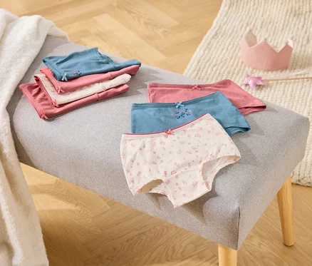 Auf einer grauen Bank liegen gefaltete Mädchenslips in Rosa, Weiß und Blau, daneben eine Spielzeugkrone und ein Zauberstab.