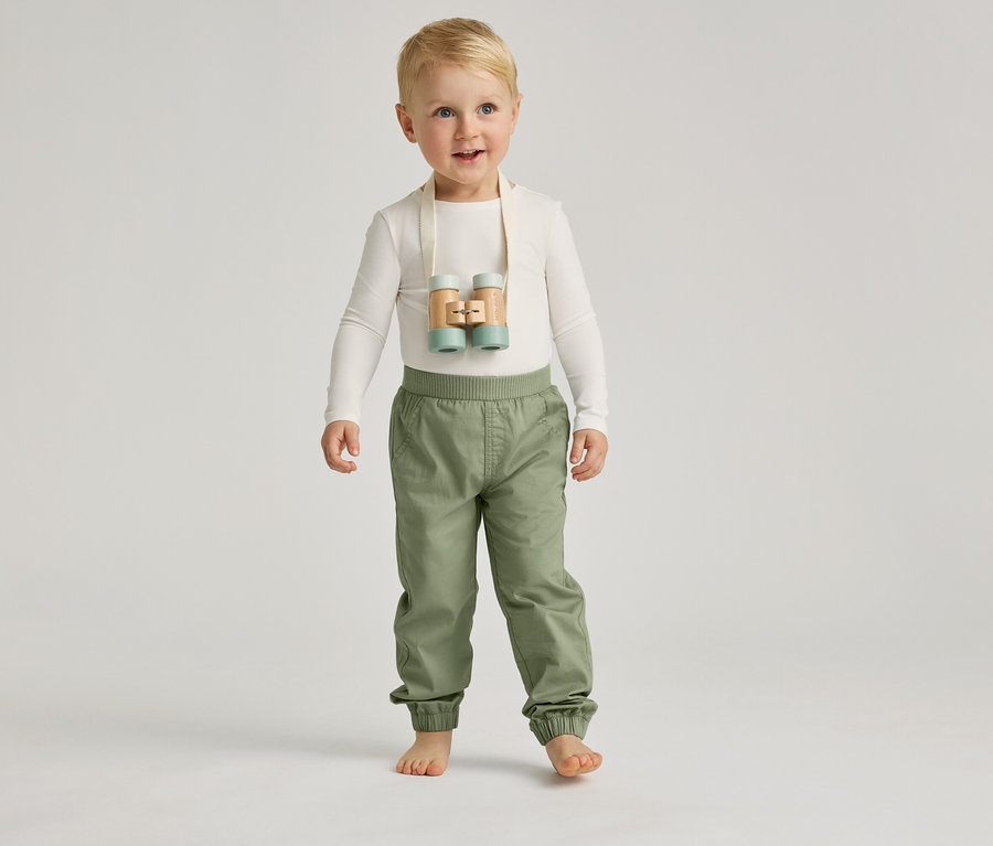 Ein barfüßiger Junge mit blauen Augen und blonden Haaren steht mit einer hellen Hose und einem hellen Oberteil vor einem grauen Hintergrund.