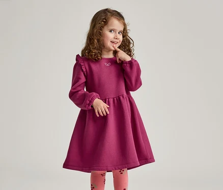 Mädchen mit lockigem Haar trägt ein weinrotes Kleid mit Rüschen und gemusterte Strumpfhosen, während sie nachdenklich ihren Finger berührt.