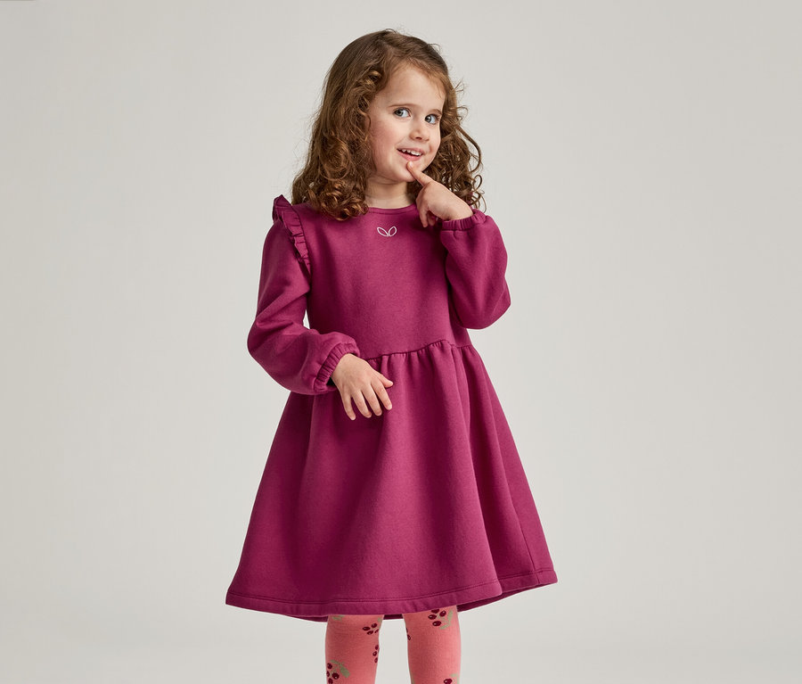 Mädchen mit lockigem Haar trägt ein weinrotes Kleid mit Rüschen und gemusterte Strumpfhosen, während sie nachdenklich ihren Finger berührt.