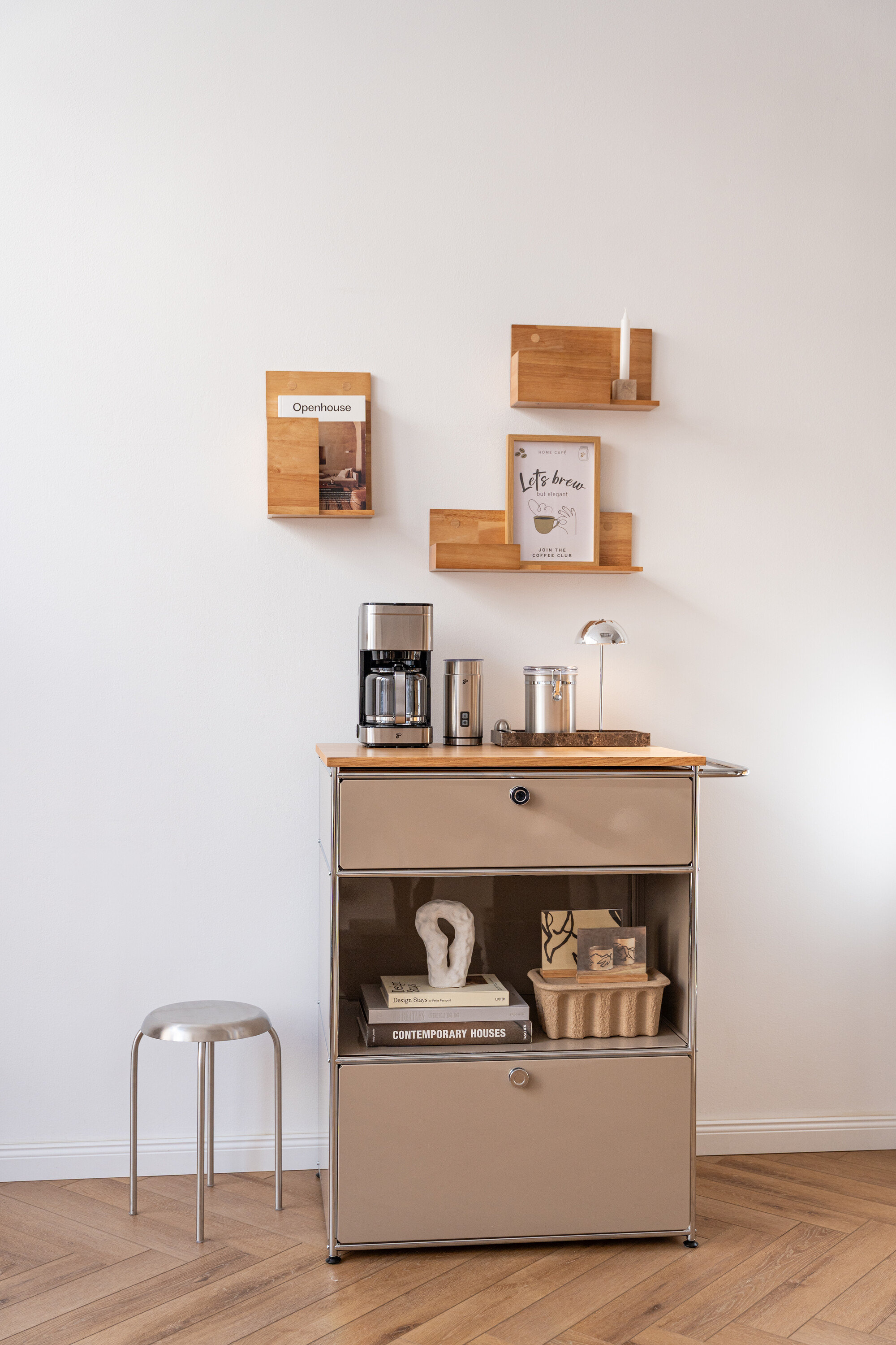 Auf einem beigen Rollcontainer stehen eine Kaffeemaschine, ein Behälter und eine Lampe. An der Wand hängen Regale mit Deko und eine Zeitschrift.