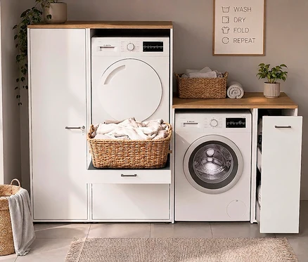 Waschküche mit gestapeltem weißem Trockner und Waschmaschine links, weiterer Waschmaschine rechts, in weißen Schränken mit Holzplatten. Wäschekörbe, Pflanzen, "WASH DRY FOLD REPEAT" Schild.