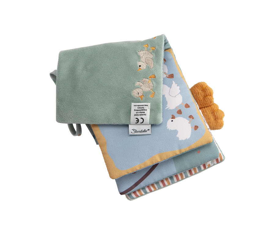 Ein teilweise geöffnetes, weiches Babybuch mit einer hellgrünen Oberseite, bestickt mit zwei Entenküken. Eine blaue Seite mit Gänsen und ein orangefarbenes Stoffelement sind sichtbar.
