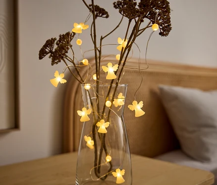 Eine klare Glasvase mit trockenen Ästen und einer Lichterkette mit leuchtenden Engelsfiguren steht auf einem Holztisch. Im Hintergrund ist ein Bett zu sehen.