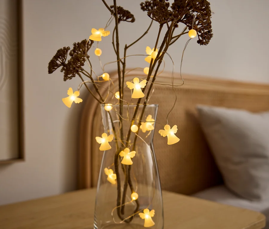 Eine klare Glasvase mit trockenen Ästen und einer Lichterkette mit leuchtenden Engelsfiguren steht auf einem Holztisch. Im Hintergrund ist ein Bett zu sehen.