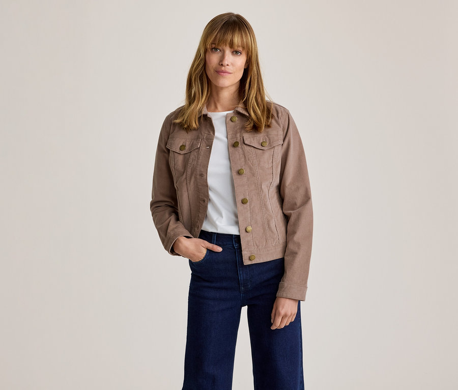 Frau mit brauner Cordjacke mit Messingknöpfen über einem weißen T-Shirt und dunkelblauen weiten Jeans, steht mit der linken Hand in der Tasche.