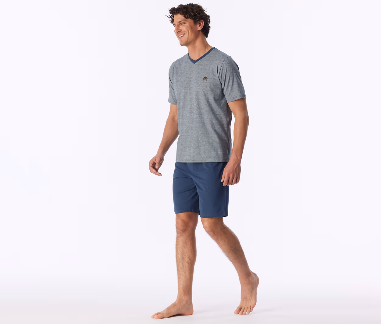 Ein lächelnder Mann mit lockigem braunem Haar geht barfuß und trägt ein graues V-Ausschnitt-T-Shirt mit kleinem Emblem und marineblauen Shorts.