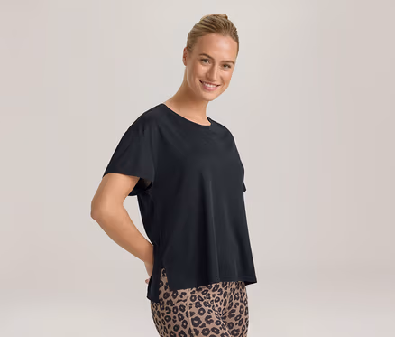 Lächelnde Frau mit Dutt, die ein schwarzes T-Shirt und Leggings mit Leopardenmuster trägt, Hände hinter dem Rücken.