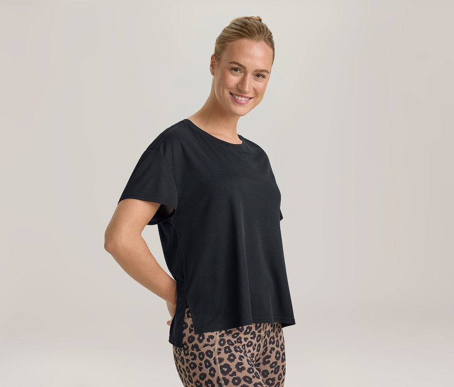 Lächelnde Frau mit Dutt, die ein schwarzes T-Shirt und Leggings mit Leopardenmuster trägt, Hände hinter dem Rücken.