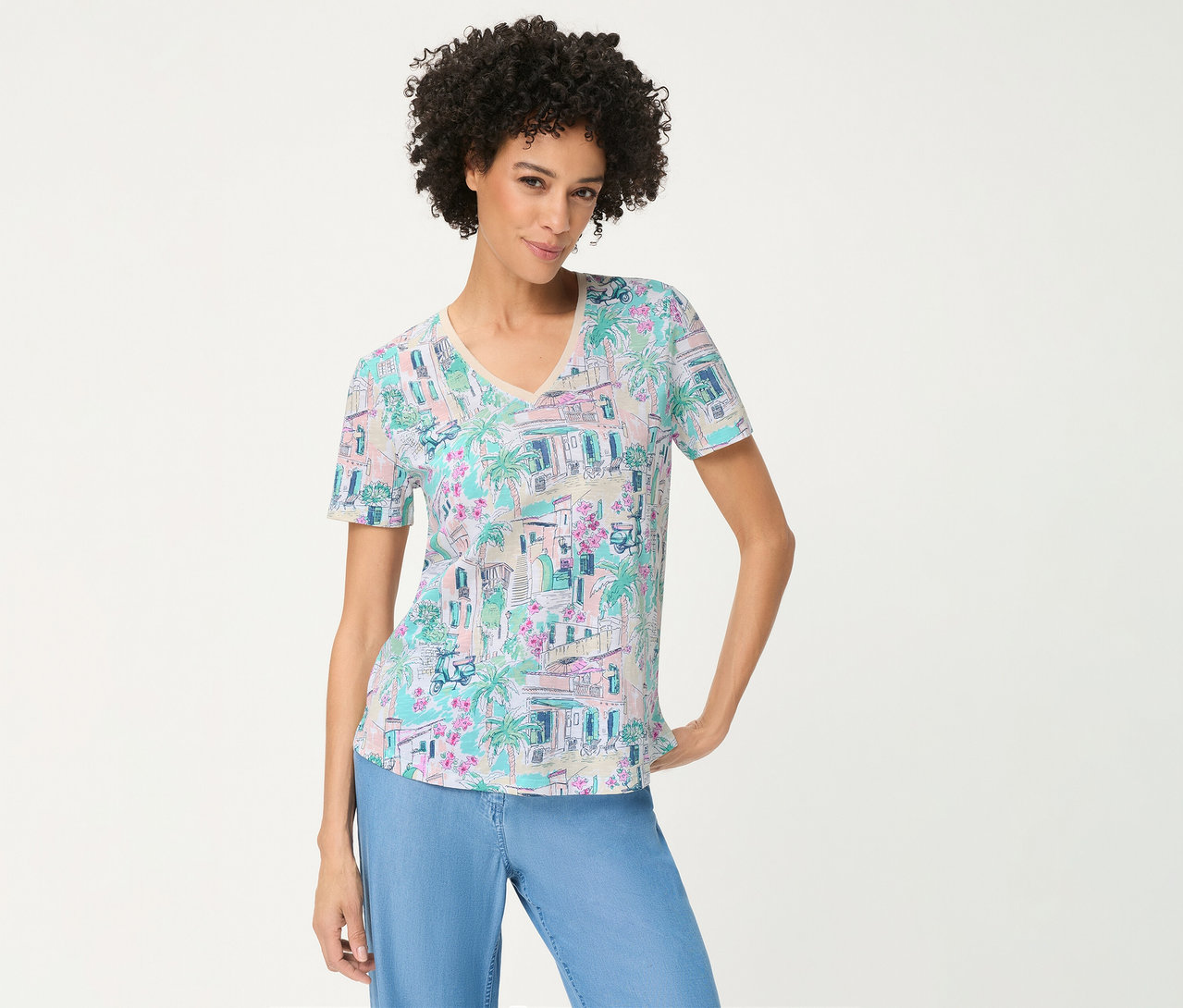 Frau mit Locken trägt ein Olsen T-Shirt mit pastellfarbenem Druck aus Gebäuden, Palmen und Blumen, dazu hellblaue Hosen, lächelt mit einer Hand an der Hüfte.