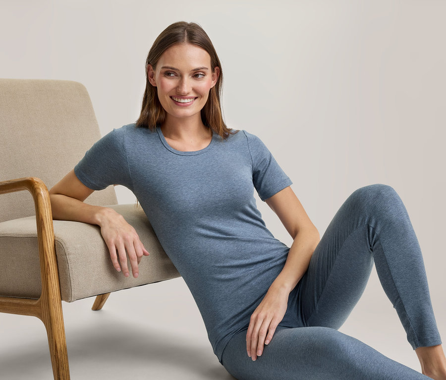 Eine lächelnde Frau sitzt auf dem Boden, lehnt an einem beigefarbenen Sessel und trägt ein blaues T-Shirt und passende Leggings.