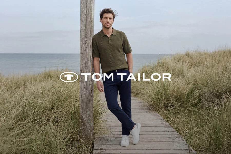 Mann in grünem Poloshirt, dunkelblauer Hose und weißen Sneakern lehnt an einem Holzpfahl auf einem Holzsteg am Meer, umgeben von hohem Gras. TOM TAILOR Logo.