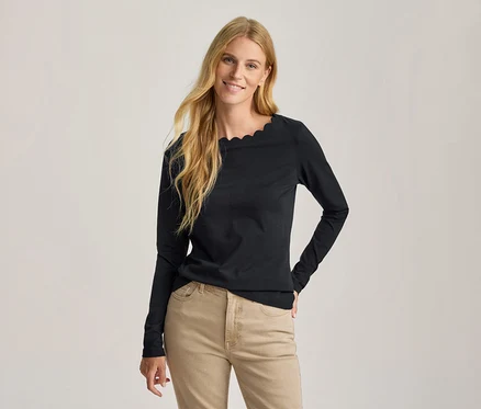 Lächelnde Frau mit langen blonden Haaren trägt ein schwarzes Langarmshirt mit Wellenausschnitt und hellbraune Hose, eine Hand in die Hüfte gestützt.