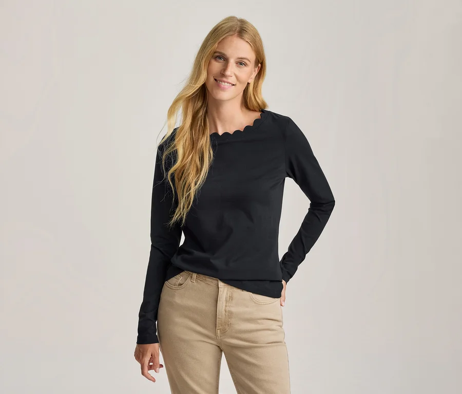 Lächelnde Frau mit langen blonden Haaren trägt ein schwarzes Langarmshirt mit Wellenausschnitt und hellbraune Hose, eine Hand in die Hüfte gestützt.