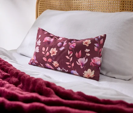 Ein Bett mit weißen Laken, einem weißen Kissen und einem rechteckigen burgunderroten Kissen mit Blumenmuster. Eine strukturierte burgunderrote Decke liegt auf dem Bett, ein Rattan-Kopfteil ist sichtbar.