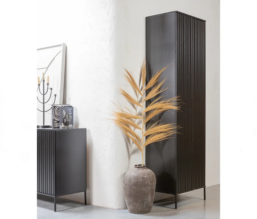 Ein Zimmereck mit einem hohen schwarzen gerippten Schrank und einem kürzeren passenden Schrank. Dazwischen steht eine große graue Vase mit getrockneten Palmblättern. Auf dem kürzeren Schrank befinden sich ein schwarzer Kerzenständer und Bücher.