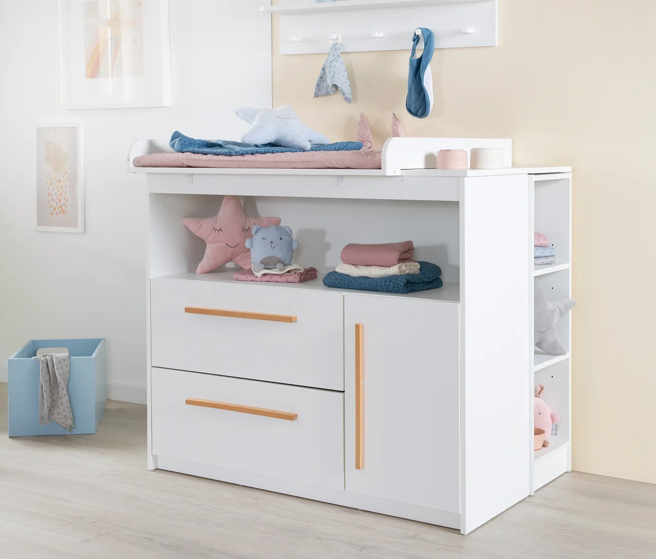 Weißer Wickeltisch des Roba 2-teiligen Kinderzimmer-Sets 2 »Lilo« mit Wickelauflage, blauer Decke und Sternenkissen. Darunter offene Fächer, zwei Schubladen und ein Schrankfach mit Holzgriffen, gefüllt mit Textilien und Stofftieren.