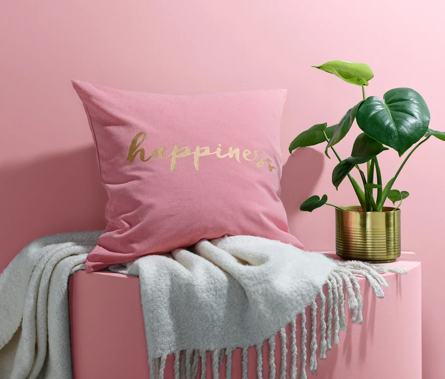 Ein rosa Dekokissen-Bezug mit dem Schriftzug Happiness liegt auf einer weißen Decke neben einer Pflanze in einem goldenen Topf.