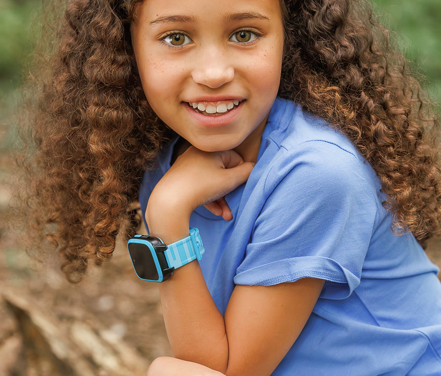 Lächelndes Mädchen mit lockigem Haar und blauer Kids Watch Xplora XGO3 2nd Gen. am Handgelenk.