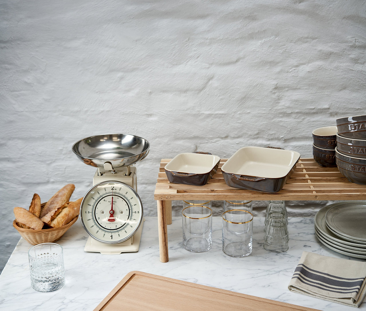 Küchentheke mit Brotkorb, Küchenwaage und zwei antikgrauen STAUB Auflaufformen auf einem Holzständer. Gläser, Schüsseln und Teller sind ebenfalls zu sehen.