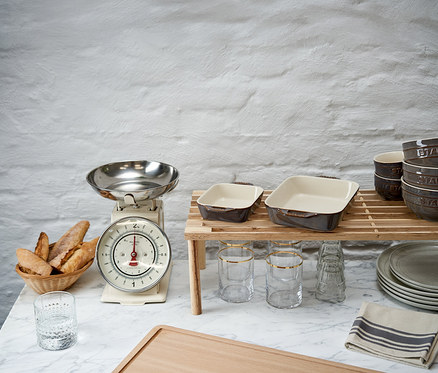 Küchentheke mit Brotkorb, Küchenwaage und zwei antikgrauen STAUB Auflaufformen auf einem Holzständer. Gläser, Schüsseln und Teller sind ebenfalls zu sehen.