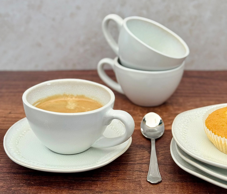 Eine gefüllte Tasse des weißen Mäser Kaffeetassen-Sets »Frozen« steht auf einer Untertasse. Daneben sind zwei gestapelte leere Tassen, ein Löffel und ein Muffin auf Tellern auf einem Holztisch zu sehen.