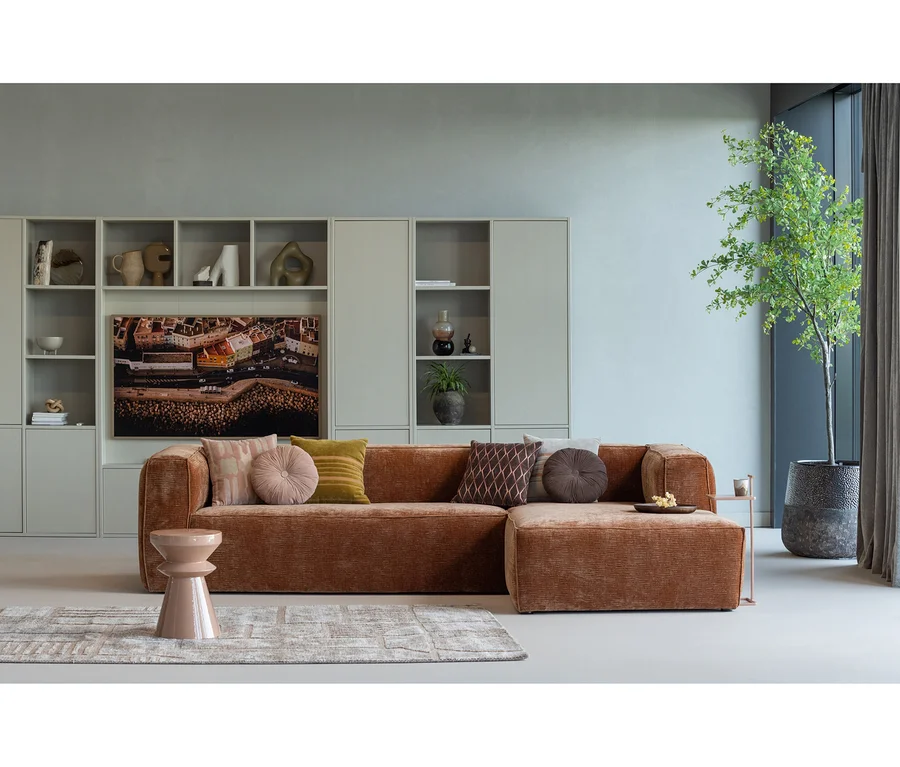 L-förmiges, rostfarbenes Sofa mit Kissen, hellem Teppich und rosa Hocker in einem modernen Wohnzimmer. Dahinter eine hellgraue Schrankwand mit Deko und großem Bildschirm, rechts eine hohe Pflanze.